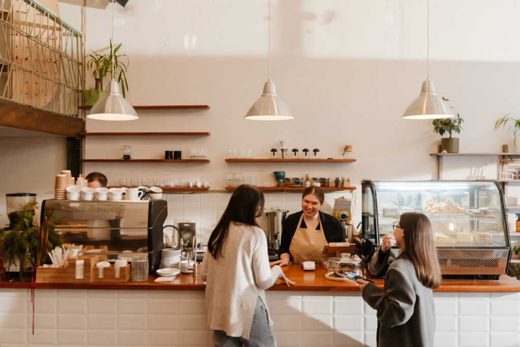 Smiling café owner helps two customers at the counter while one taps to pay at the point of sale terminal