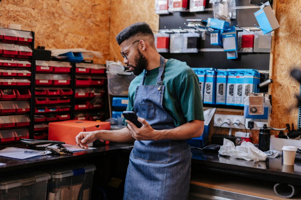 Small business owner in an apron checking his phone in his repair shop, reviewing merchant processing fees and transactions