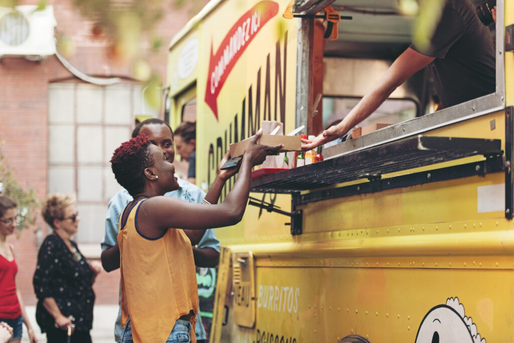 Customer receives her order at a food truck while paying with her mobile phone, as other customers wait in line