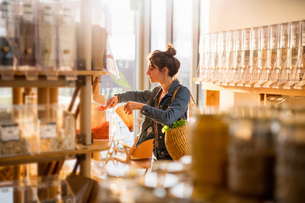 10 Tips to Create the Best Retail Checkout Experience 1 A woman with a woven basket bag fills a paper bag from bulk bins of grains and dried goods in a sunlit refill store, browsing at her own pace before checkout.