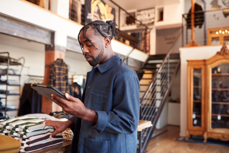 A small business owner reviews payments on a tablet while working the floor of his clothing shop