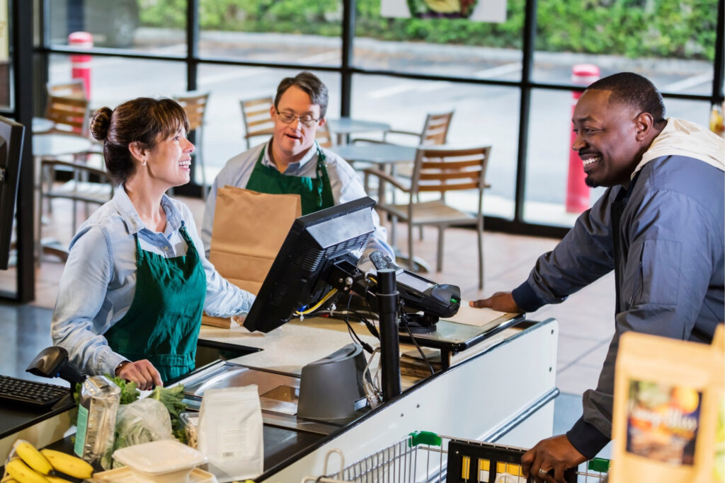 10 Tips to Create the Best Retail Checkout Experience 2 A smiling cashier and a bagger with Down syndrome help a customer at a grocery checkout counter, showing how well-staffed, friendly service shapes the checkout experience.