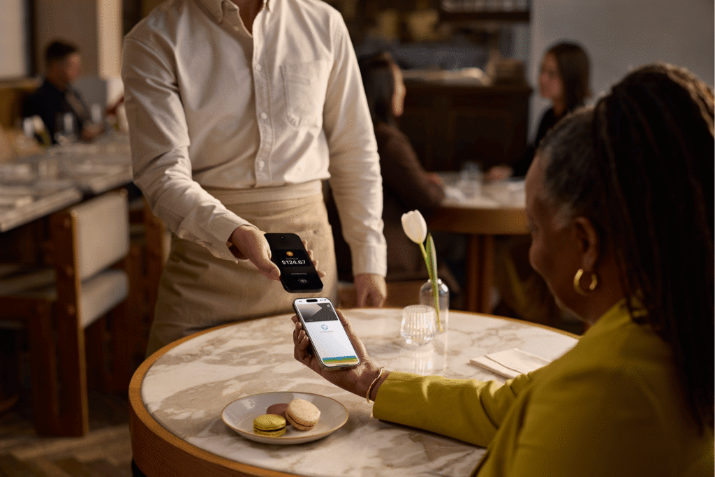A diner holds her phone beneath a server's smartphone to pay a $124.67 restaurant bill using tap to pay.